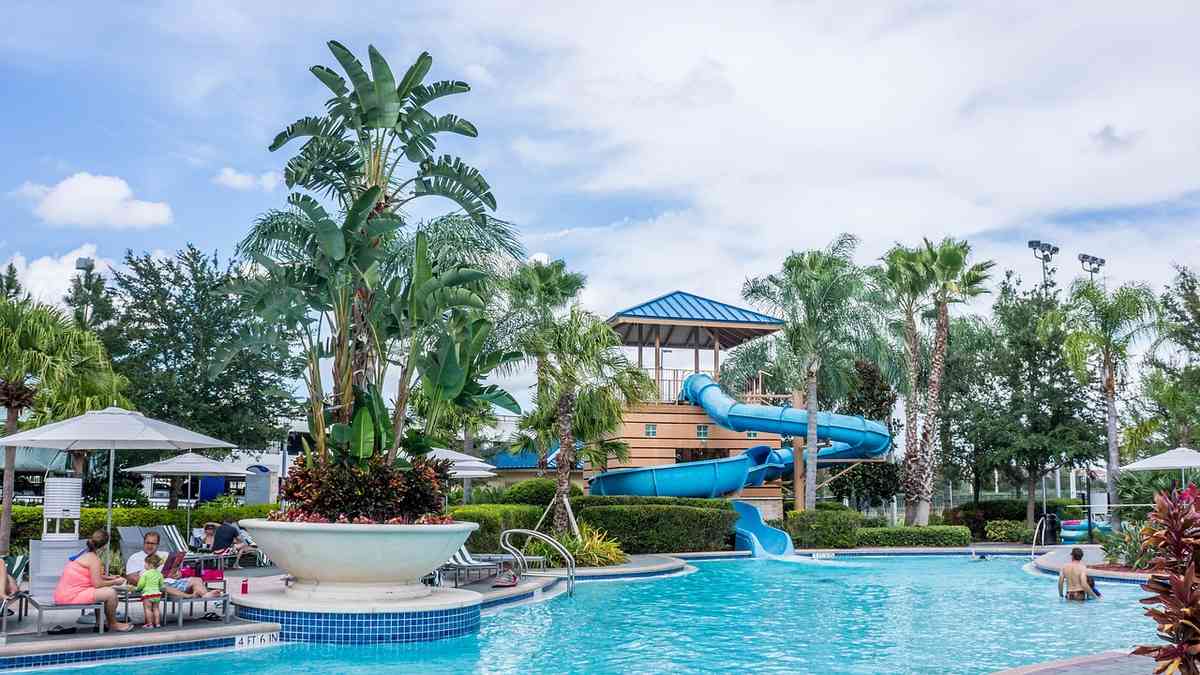 Piscine avec toboggan bleu entouré de palmiers et de parasols sous un ciel nuageux Piscine avec toboggan bleu entouré de palmiers et de parasols sous un ciel nuageux