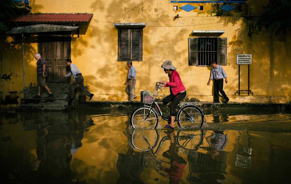 Femme sur vélo devant maison jaune, eau stagnante, personnes marchant, clôture, végétation Femme sur vélo devant maison jaune, eau stagnante, personnes marchant, clôture, végétation